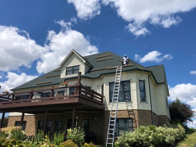 A worker on the roof of a large two-story house with green shingles and beige siding. The house has a deck and stone foundation, with landscaping in the foreground. A ladder is propped against the house, reaching the second story under clear blue skies.